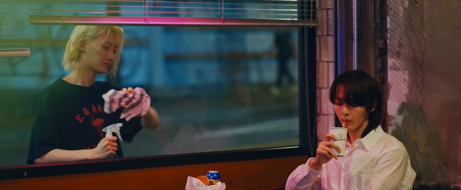 still from YooJung's "begin" MV. KB sits inside and looks down at what he's drinking. YooJung stares at him from outside the window as he washes it with a rag.