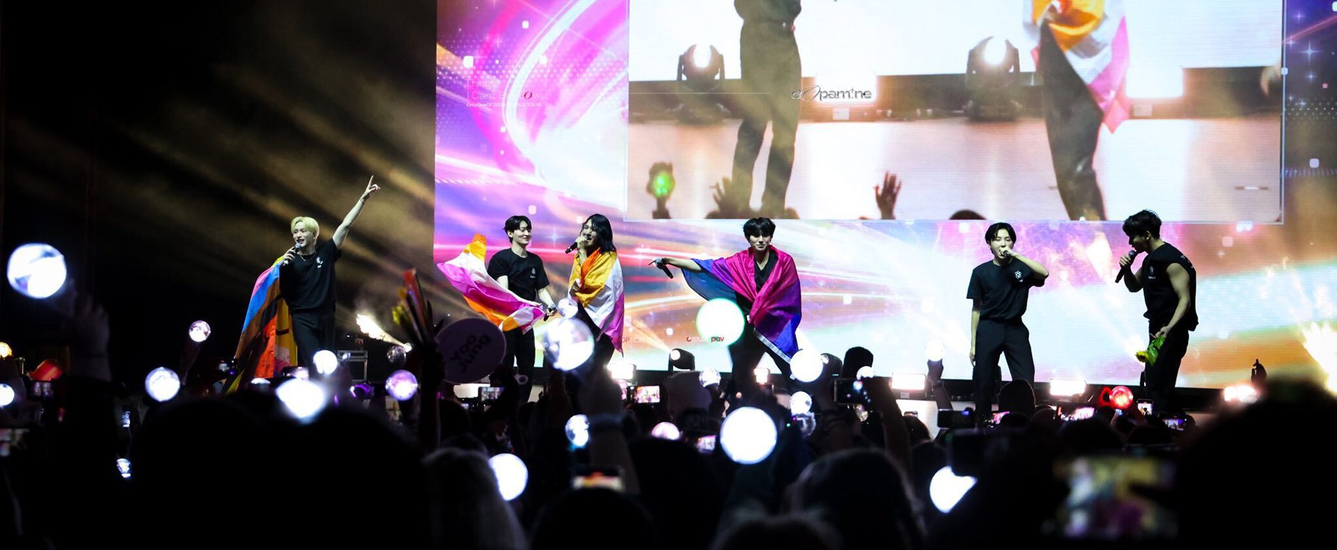 a photo taken during the New York stop of the Things I Can't Say LOve tour. the OnlyOneOf members stand on stage from left to right: Nine, YooJung, JunJi, Mill, Rie, KB. Nine holds up the Philly pride flag while YooJung holds the lesbian pride flag. JunJi is wrapped in a lesbian pride flag; Mill is wrapped in a bisexual pride flag.