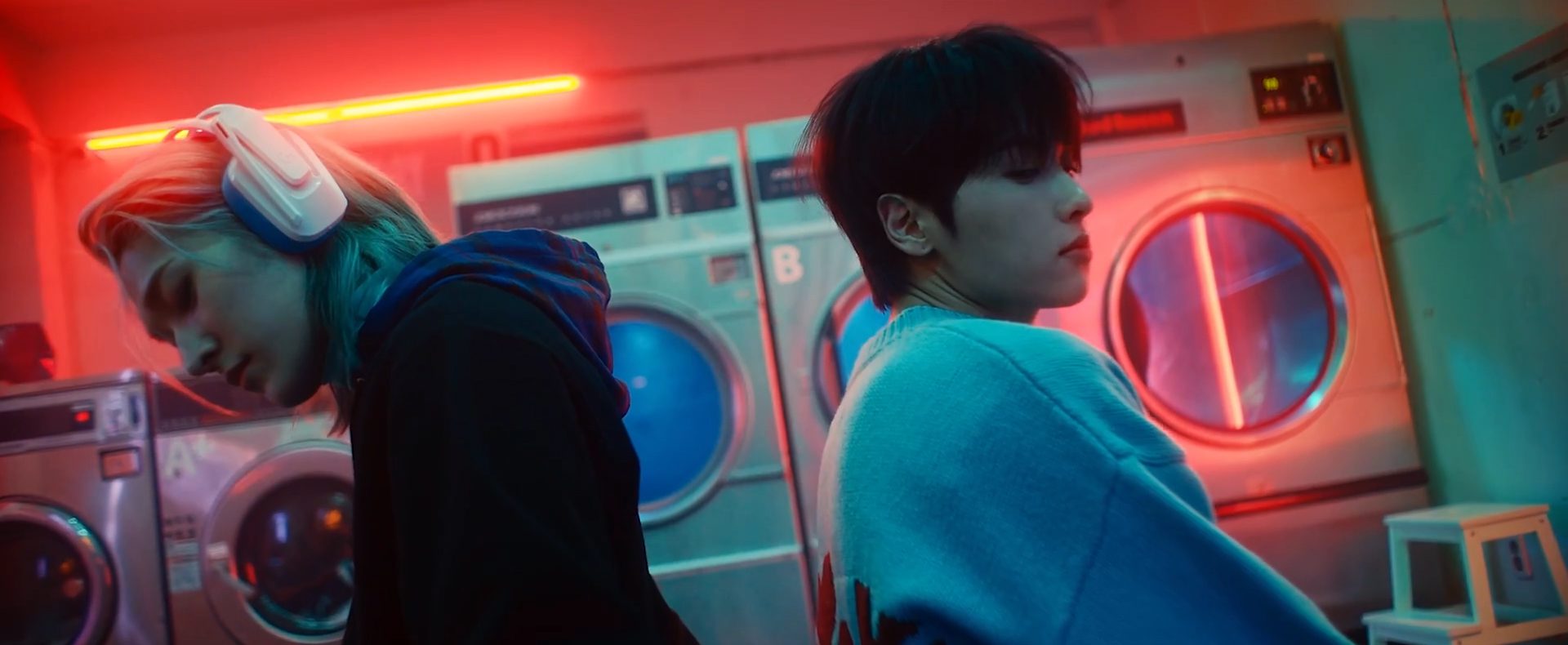 still from OnlyOneOf's "seOul drift" MV. JunJi and Rie sit back-to-back in a laundromat. JunJi is wearing headphones. the pair look down, heads slightly turned towards the camera.