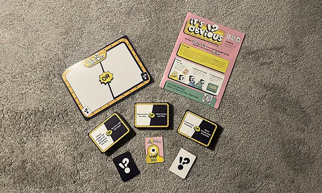 The contents of It's Obvious are laid out on top of a grey carpet. The instructions are on top next to the custom question board. Three stacks of question cards are in the middle. 