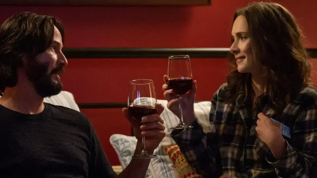 Keanu Reeves and Winona Ryder hold wine glasses, looking at each other and sitting in a bed together. 