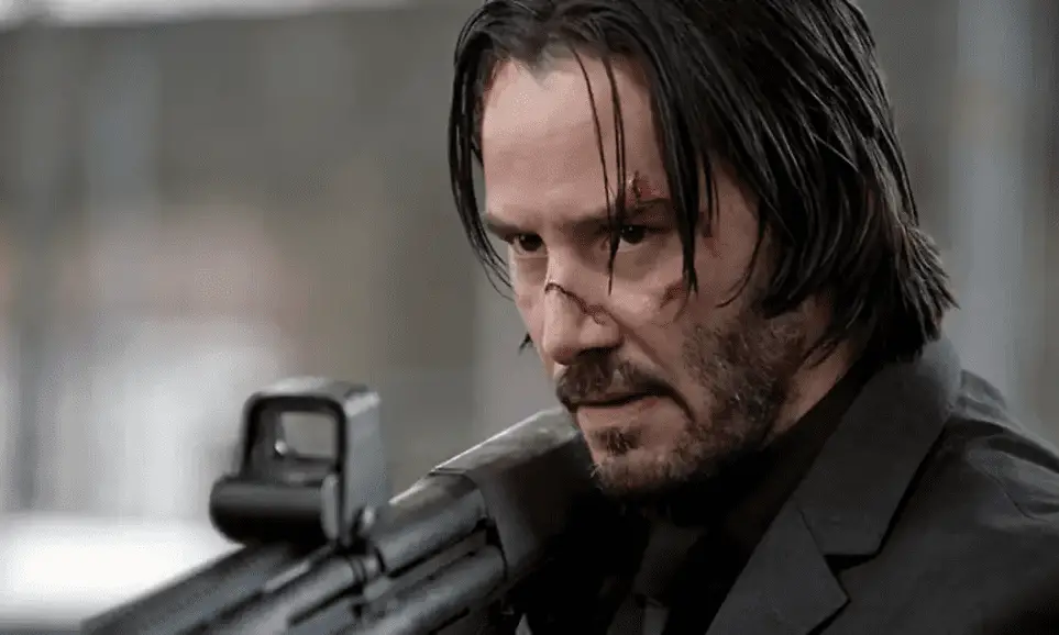 Keanu Reeves as John Wick with scars on his face, holds a shotgun in this close up