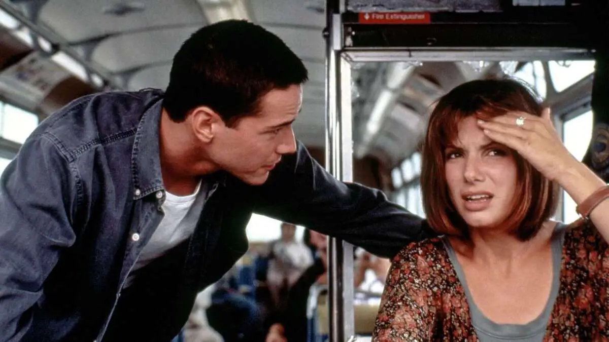 Sandra Bullock sits in the driver's seat of a bus as she puts her hand on her forehead, and Keanu Reeves leans in with his hand on the back of her chair. 