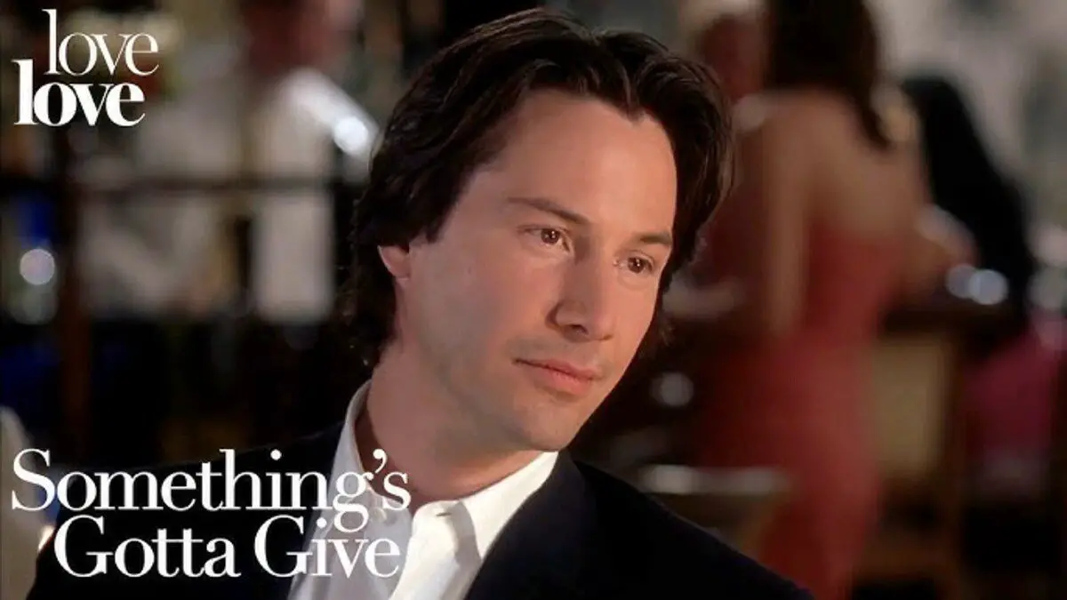 Keanu is sat in a restaurant with the background out of focus as he looks forward and to the right. 