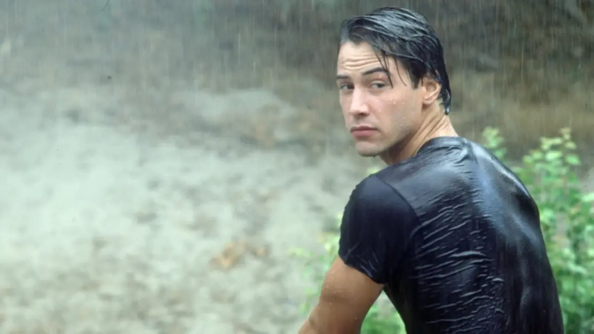 Keanu Reeves faces his back away from the camera, and turns his head to look back towards the camera in the rain, outside. 