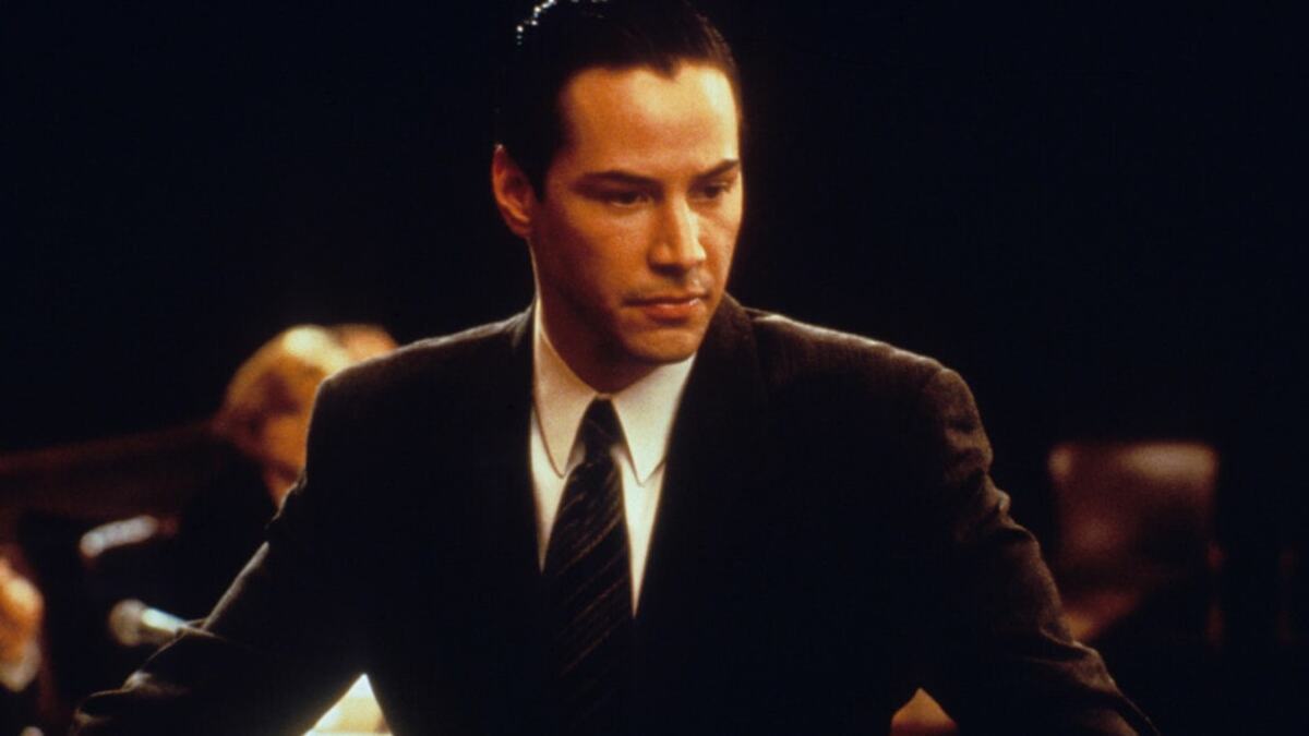 Keanu Reeves wears a suit in a dark courtroom. 